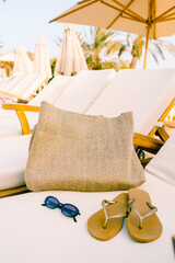 Close-up of a raffia beach bag, flip-flops, and sunglasses on a sun lounger under a parasol. Natural textures and warm tones evoke a relaxed, sustainable summer lifestyle.