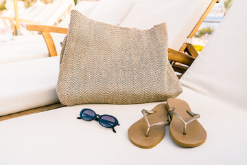A raffia tote beach bag, sunglasses, and flip flops rest on a clean white lounger with a glimpse of water between chairs. A stylish and tranquil summer setup at a resort by the sea or pool.
