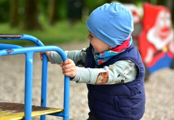Obraz premium Smiling toddler boy playing at a playground, holding on to colorful bars. Dressed warmly in a beanie and vest, he enjoys outdoor playtime on a chilly day surrounded by nature.