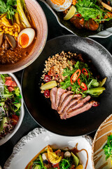 A variety of gourmet dishes are beautifully presented, including a bowl of ramen with a soft-boiled egg, a fresh salad with greens and tomatoes, and sliced meat garnished with herbs, nuts, and pickles