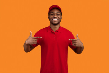 Fototapeta premium Choose Me. Smiling Bearded African American Delivery Man Pointing Fingers At Himself And At Red T-shirt Advertising Courier Delivering Service Posing Over Yellow Background. Studio Shot