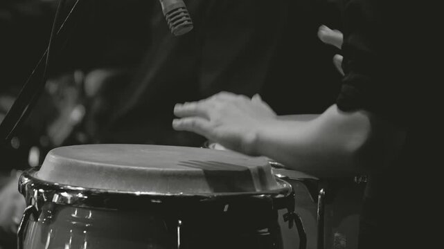 Close-up of conga drums, showing the rhythm and movements of the musician's hands. In the background, a blurred drummer playing the drums. Black and white footage