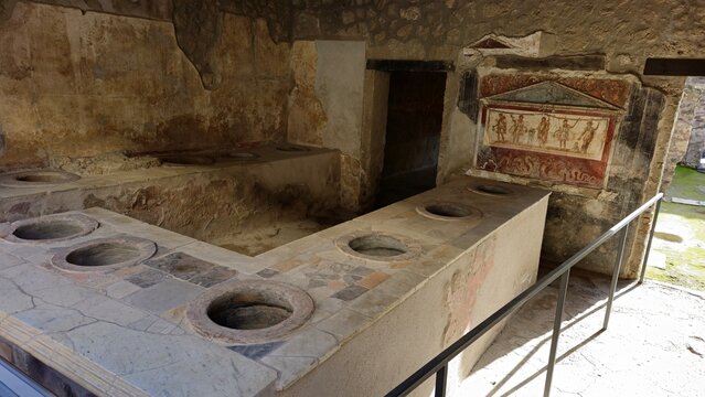Pompeii, Italy - 8 January 2025. Interior of a thermopolium features stone counters with embedded dolia and a fresco of five figures above a decorative frieze on the back wall.