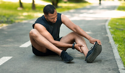 Sports injury. African American runner sitting on jogging track and feeling pain in his ankle,...
