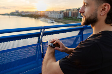 Young sporty man jogger taking break from training outdoors using smart fitness watch and looking...