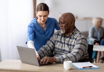 Friendly young woman nurse helping black senior man in weheelchair using laptop, smiling elderly...