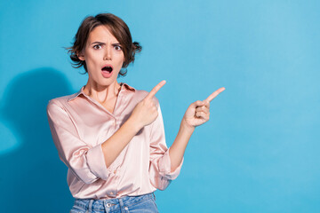 Surprised young woman in stylish pink blouse pointing sideways against a blue backdrop
