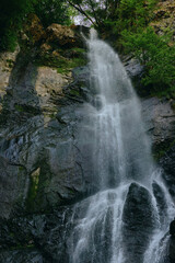 Fototapeta premium Majestic waterfall cascading over rocky cliffs surrounded by lush green forest and bright natural light illuminating the flowing water in a serene outdoor setting.