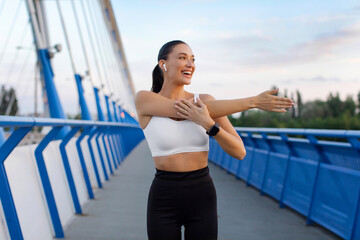 Smiling lady in sportswear warming up and listening to music in earphones, stretching her arms, doing sports outdoors on city footbridge