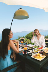 A brunette and blonde in their 20s enjoy breakfast together at a hotel table with beautiful...