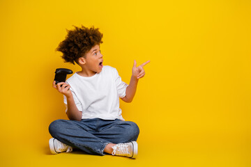 Excited young boy with curly hair holding video game controller on a bright yellow background, pointing in surprise © deagreez