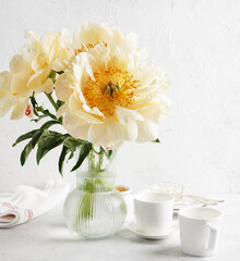 Creamy peonies in vase and cups for tea on white table in morning lights, copy space