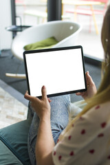 Femme assise confortablement dans un intérieur moderne, tenant une tablette numérique avec un écran blanc. Scène de détente en journée, avec lumière naturelle entrant par de grandes baies vitrées	