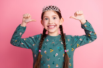 Smiling young girl with a tiara and pigtails gesturing confidently against a vibrant pink background