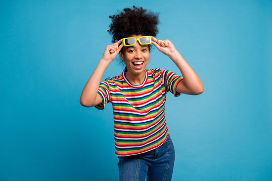 Cheerful young woman in striped shirt adjusting sunglasses on vibrant teal background