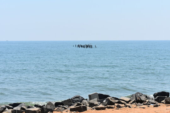 Rocky shoreline at Promenade Beach with calm blue sea and distant wooden posts on the horizon.