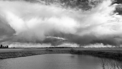 Monochrome rural landscape in the agricultural field at the dugout filled with water, farm water collection supply system. Dramatic rainy clouds in the sky.
