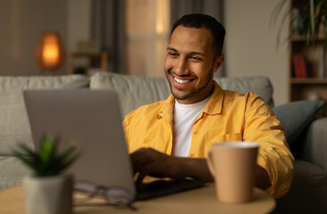 Smiling young black man working or communicating online, sitting on couch with laptop at home....