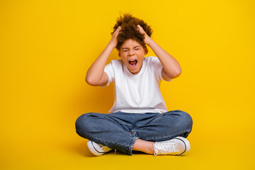Young boy expressing frustration with hands on head while sitting against a bright yellow...