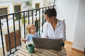 Mother Working on Laptop While Engaging With Child on Outdoor Balcony