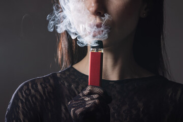 Stylish woman exhales thick vapor while holding a red electronic vape. Dressed in elegant black...