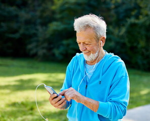 Happy active senior man having fun using smartphone and wearing earphones and sportswear, checking music or an exercise app, after having an exercise sport activity outdoors