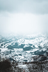 Obraz premium Skiers enjoy the slopes of Gala Yuzawa with stunning views of Ishiuchi Maruyama and Yuzawa Kogen in Niigata, Japan. Snow-covered mountains and a small town stretch across a scenic winter valley.