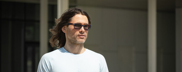 Banner of athletic caucasian man with long curly hair wearing sunglasses and light blue T-shirt walks confidently outdoors. His thoughtful expression highlights the calm and relaxed atmosphere of