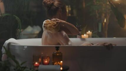 A woman with auburn hair in a bun enjoys a relaxing self-care moment, scrubbing her back with a soapy sponge in a white bathtub. Surrounded by plants and candles in a dimly lit bathroom, she finds tra - Powered by Adobe