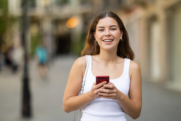Young smiling girl with phone walks along city street, walks from work to home. Pedestrian in...