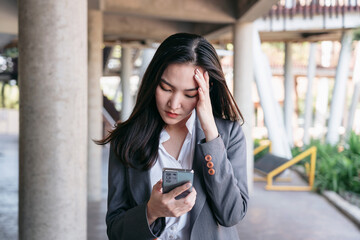 Asian businesswoman in suit, using smartphone for reply email to client and holding her head with...
