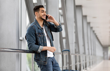 Mobile Communication. Smiling Young Arab Guy Talking On Cellphone At Airport, Millennial Middle Eastern Man Standing With Luggage Near Window At Terminal And Enjoying Pleasant Call, Copy Space