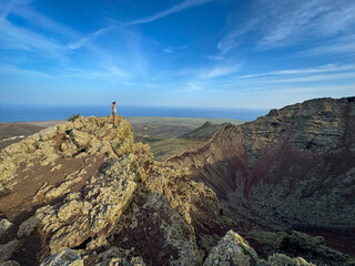 Fototapeta premium A lone female figure stands on a rocky volcanic rim, overlooking both a massive crater and the distant Atlantic Ocean. Stunning panoramic view from popular geological attraction at Lanzarote island.