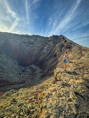 Fototapeta premium Woman stands on the rugged edge of a vast volcanic crater, gazing into its depths under a partly cloudy sky. Dramatic landscape shows immense scale and raw power of the natural geological formation.