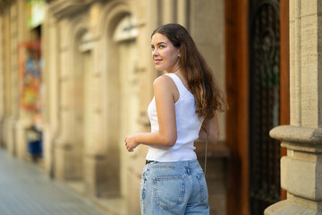 Young woman in early 20s wearing white tank top and blue jeans gently smiling and turning back...