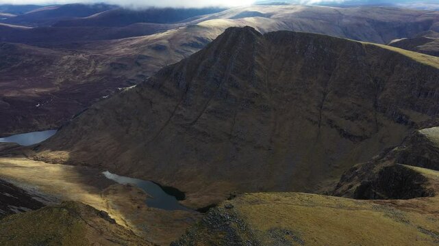 Aerial view of a hiker on the summit of the Scottish mountain Seana Bhraigh in Scotlands Highlands, overlooking the dramatic peaks, hills and cliffs as the sun breaks through the clouds