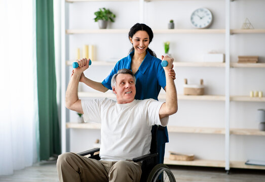 Post-stroke rehabilitation for disabled people. Millennial physiotherapist helping elderly male patient in wheelchair to make exercises at home. Impaired senior man working out with dumbbells