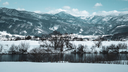 Peaceful winter landscape of snowy mountains and a calm river under a blue sky in Yuzawa, Japan. Bare trees and snow-covered fields create a serene, panoramic scene of rural natural beauty.