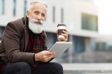 Handsome aged grey-haierd man holding paper cup with coffee and using modern digital tablet,...