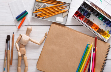 Workplace of artist. Paints, sketchbook and wooden painter man with brushes on white desk, top...