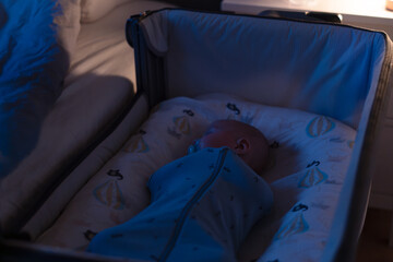 A baby wrapped snugly in cocoon swaddle bag is peacefully sleeping in a crib at night near parent bed. Dimly lit, calm atmosphere ideal for rest. Safety newborn baby sleep. Soft selective focus © okrasiuk