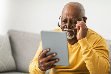 Senior Black Man With Poor Eyesight Using Digital Tablet Wearing Glasses And Squinting Eyes Reading...