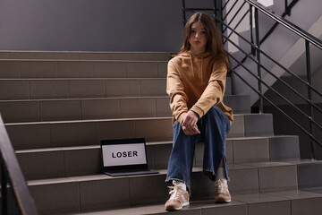 Cyber bullying. Upset teenage girl and laptop with text You Loser on stairs indoors. Space for text