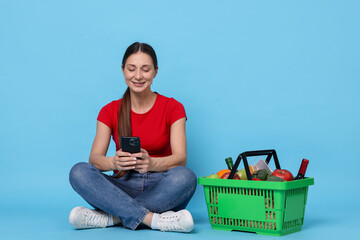 Smiling woman with basket full of products and smartphone on light blue background. Online shopping