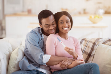 Obraz premium Young African American family learning about their future child from pregnancy test at home, embracing on sofa. Loving husband and his beautiful expectant wife cuddling and feeling happy