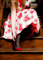 Fototapeta premium A woman in a floral dress and black stockings is sitting on wooden stairs, adjusting her red shoe.