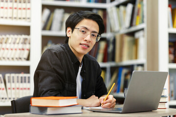 Working On Research Project. Portrait of asian male student using laptop at library, writing in...