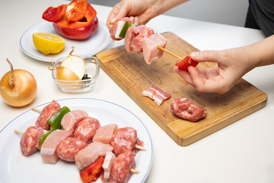 Hands of female chef preparing meat and vegetable skewers with pork, bacon, red and green bell peppers, and onions on a wooden cutting board in kitchen