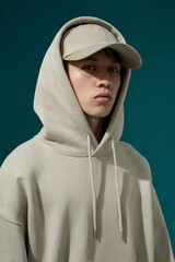 A fashion photography portrait against a deep teal colored background. The subject wears an oversized beige hooded sweatshirt with drawstrings and a matching beige baseball cap worn backwards.
