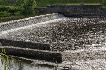 Backlit image of a weir with cascading water and sparkling reflections on a rural river. A mix of nature and engineering, calm yet dynamic